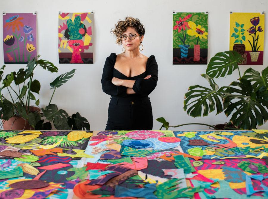 Photo of artist Favianna Rodriguez, arms crossed and in a black dress, behind a table full of her paintings and posters, and in between to potted plants.