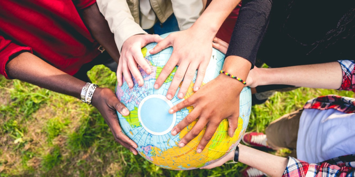 A photo from above of the arms of 4 people, of different skin colors and styles, holding a plastic world globe