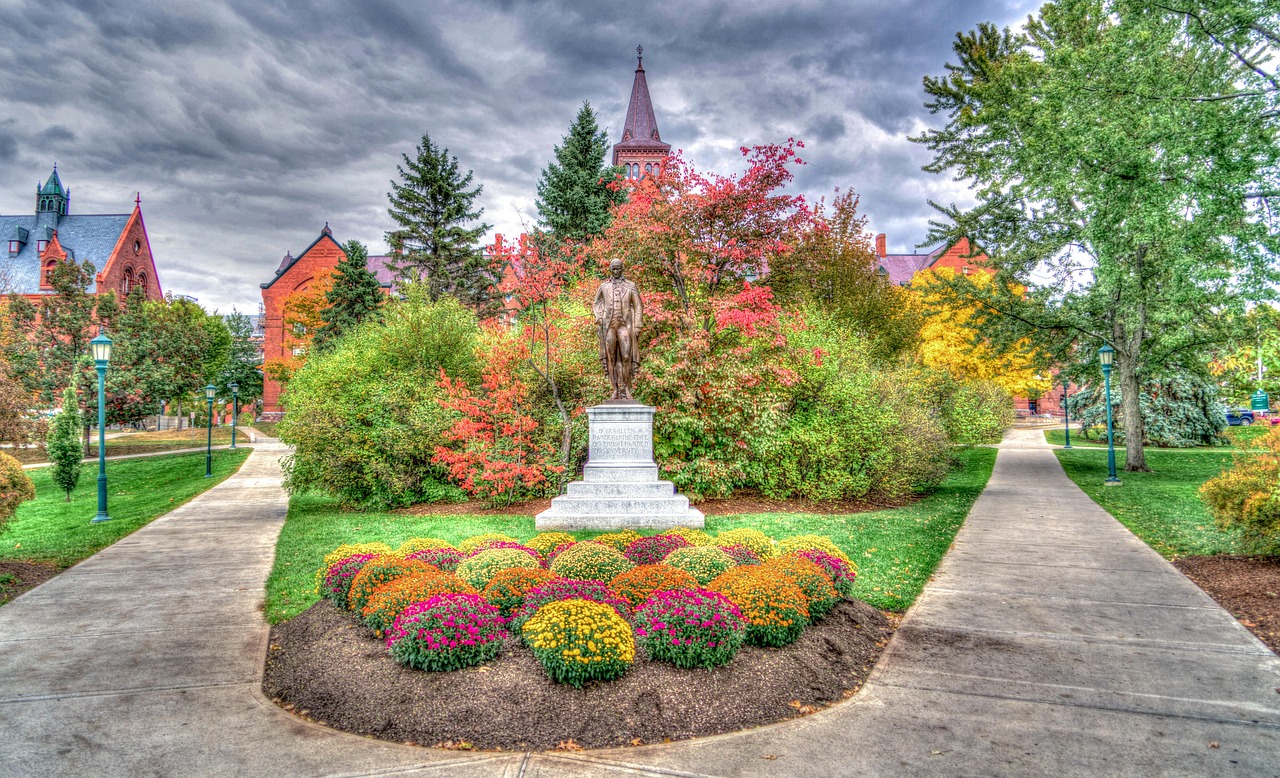Photo of entrance to University of Vermont, two concrete pathways in the middle flower beds with a lot of colors and a bronze statue in the middle. In the background a Victorian type of building with a bell tower
